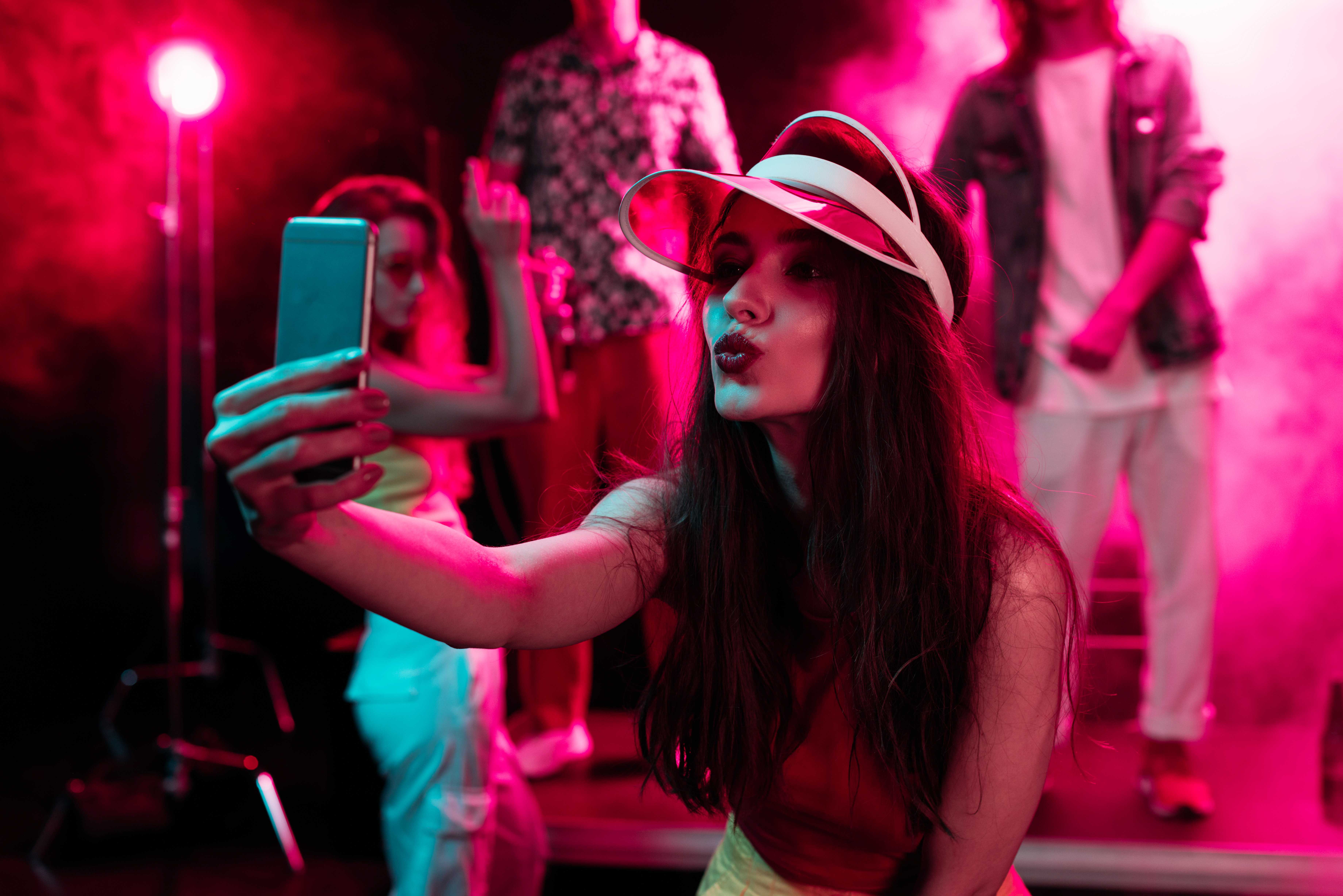 A young woman in a visor poses for a selfie in a room lit by pink neon lights. Behind her are another young woman and two men dancing surrounded by smoke